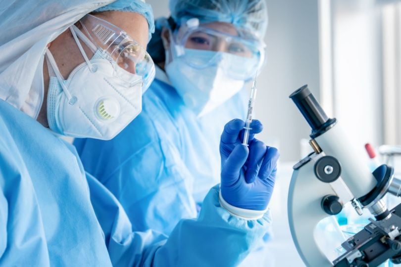 Two healthcare professionals in protective gear with a syringe and a microscope.