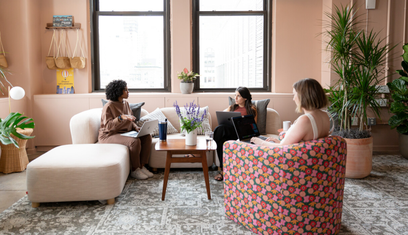 Three monday.com team members sit on upholstered chairs with laptops, talking 