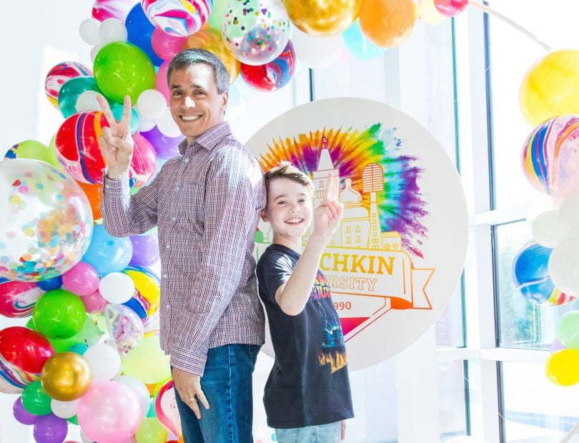 Munchkin team member stands back to back with his son in front of Munchkin University sign and balloons.