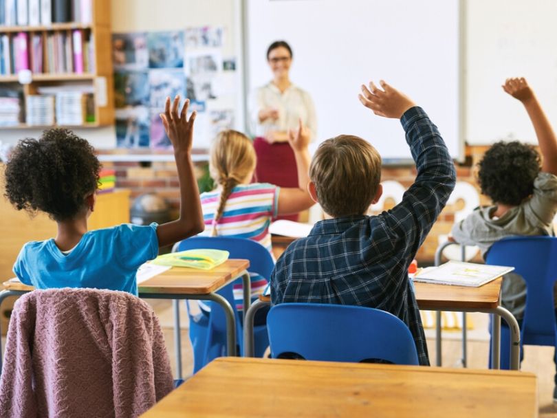 Children seated at desk raise hands to answer question posed by teacher standing in front of whiteboard