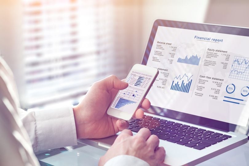 A use sits in front of a laptop while holding his phone; both display graphed financial data