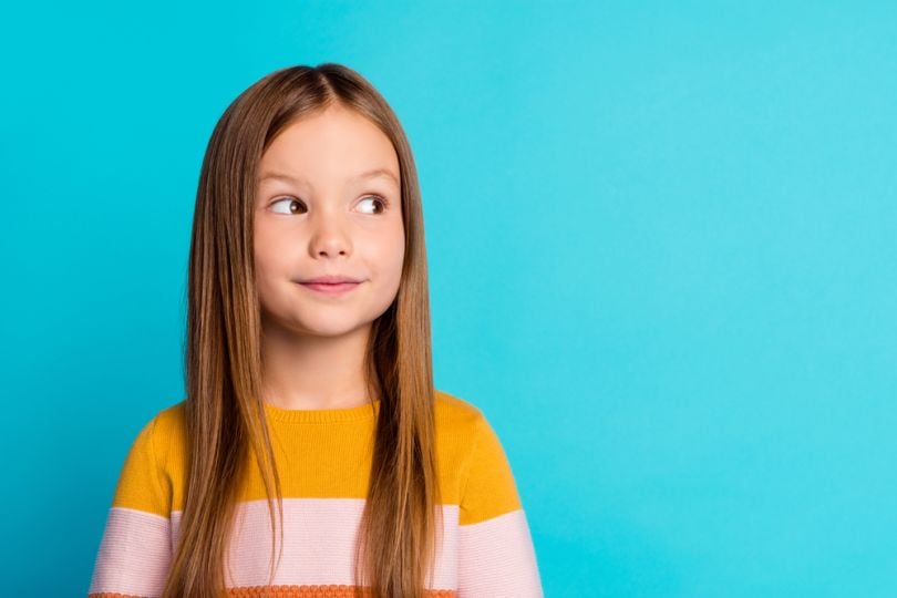  Child with long hair against a blue backdrop, looking to the side with a slight smile.