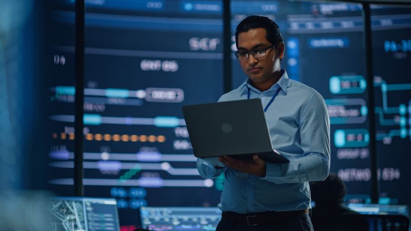 Cybersecurity professional holding laptop while standing inside monitoring a security operations center.