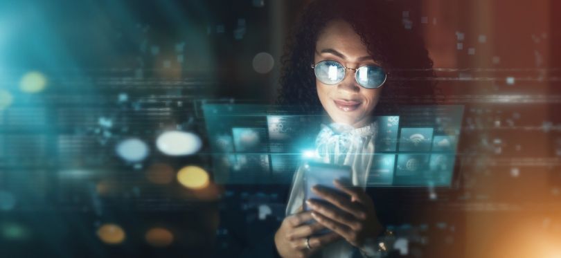 A woman wearing glasses looking down at her phone in a dark room, with her lenses and a glass pane in front of her reflecting digital screens. 