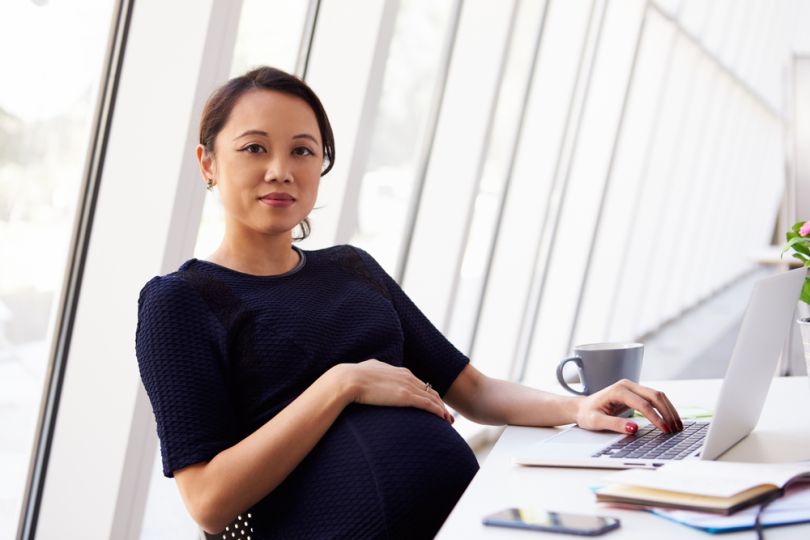 Pregnant Asian woman sits back at desk, one hand over her belly, the other hand on the keyboard of a laptop. 