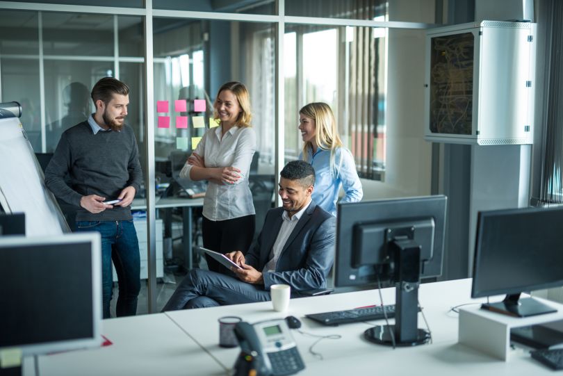 Group of co-workers having a standing huddle near a glass post-it wall and computers