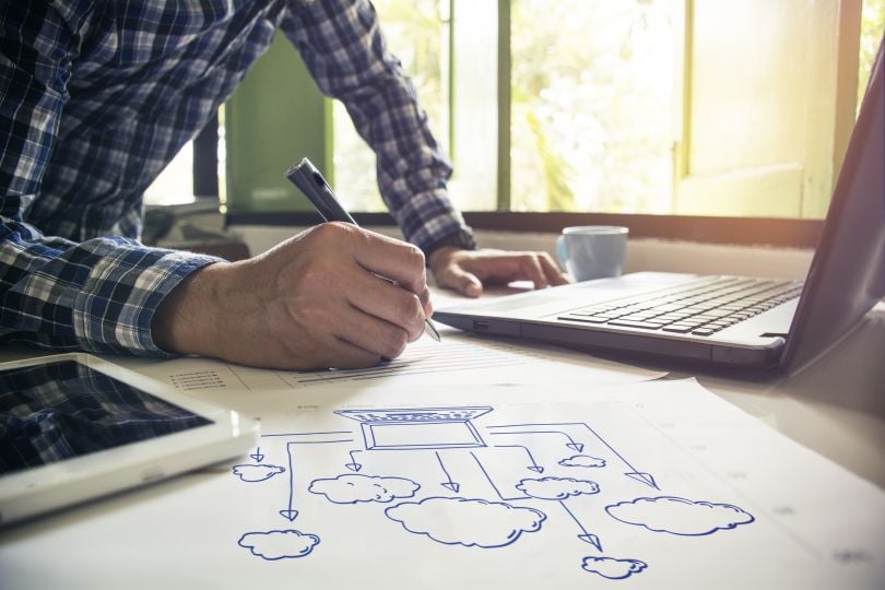 An office worker sketches a physical blueprint abstracting the concept of cloud computing while referencing a laptop