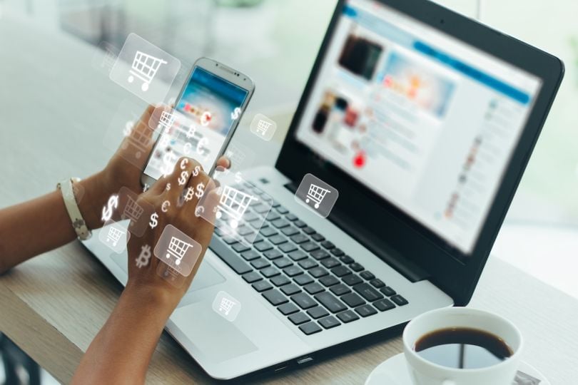 A person using a smartphone with shopping and money transfer icons floating around it in front of an open laptop.