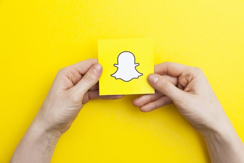 Two hands holding a sticky note with the Snapchat ghost logo on a bright yellow background.