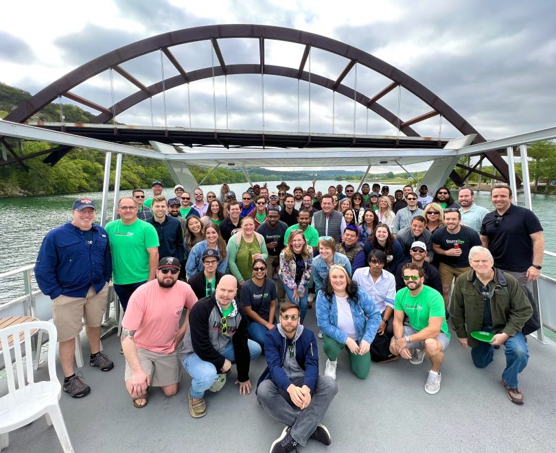  Large group photo of SourceDay team members on a ferry with a semicircular bridge in the background.