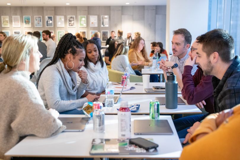 A diverse group of Sprout Social employees gather around a cluster of tables amid a crowd of peers also gathered at tables in the background.