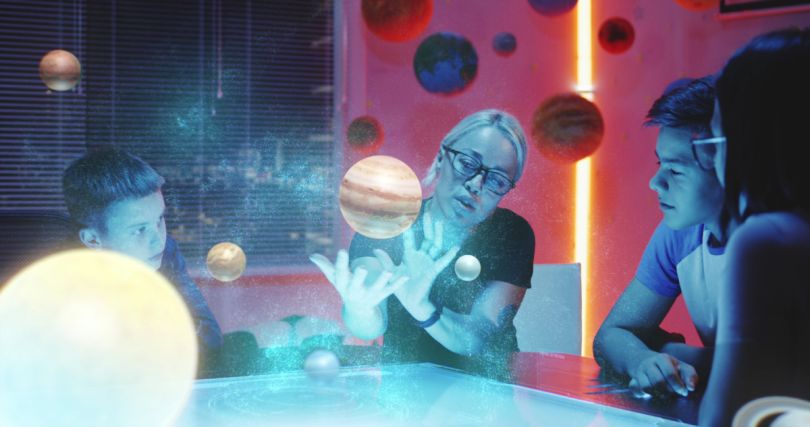 A woman motions toward holograms of planets while children watch. 