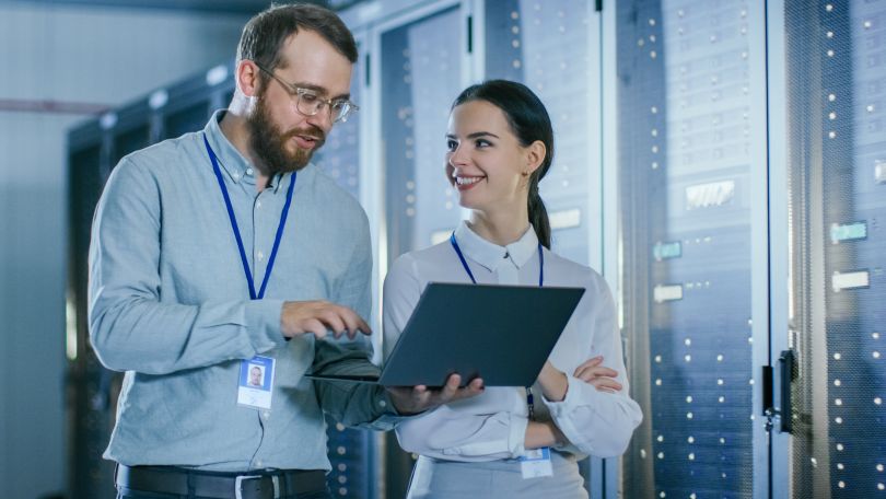Image of an employee holding a laptop in an IT setting, talking to a coworker standing beside them